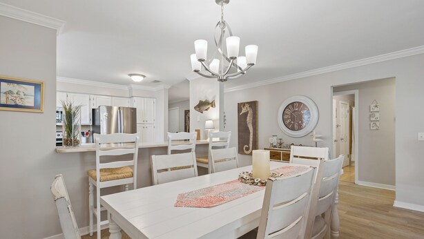 Un coin repas avec une table blanche, des chaises, un lustre et une cuisine en arrière-plan.