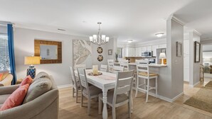 Un coin repas avec une table et des chaises blanches, un lustre et une cuisine en arrière-plan.