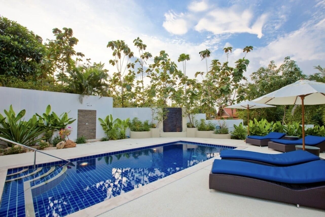 Una piscina con baldosas azules, rodeada de sillas de descanso y sombrillas, con una pared blanca y follaje al fondo.