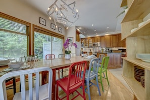 A modern kitchen with wooden cabinets, a dining table with chairs, and a view of the outdoors through large windows.