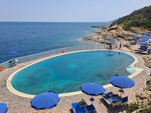 A clear blue pool surrounded by stone tiles, with blue umbrellas and lounge chairs.