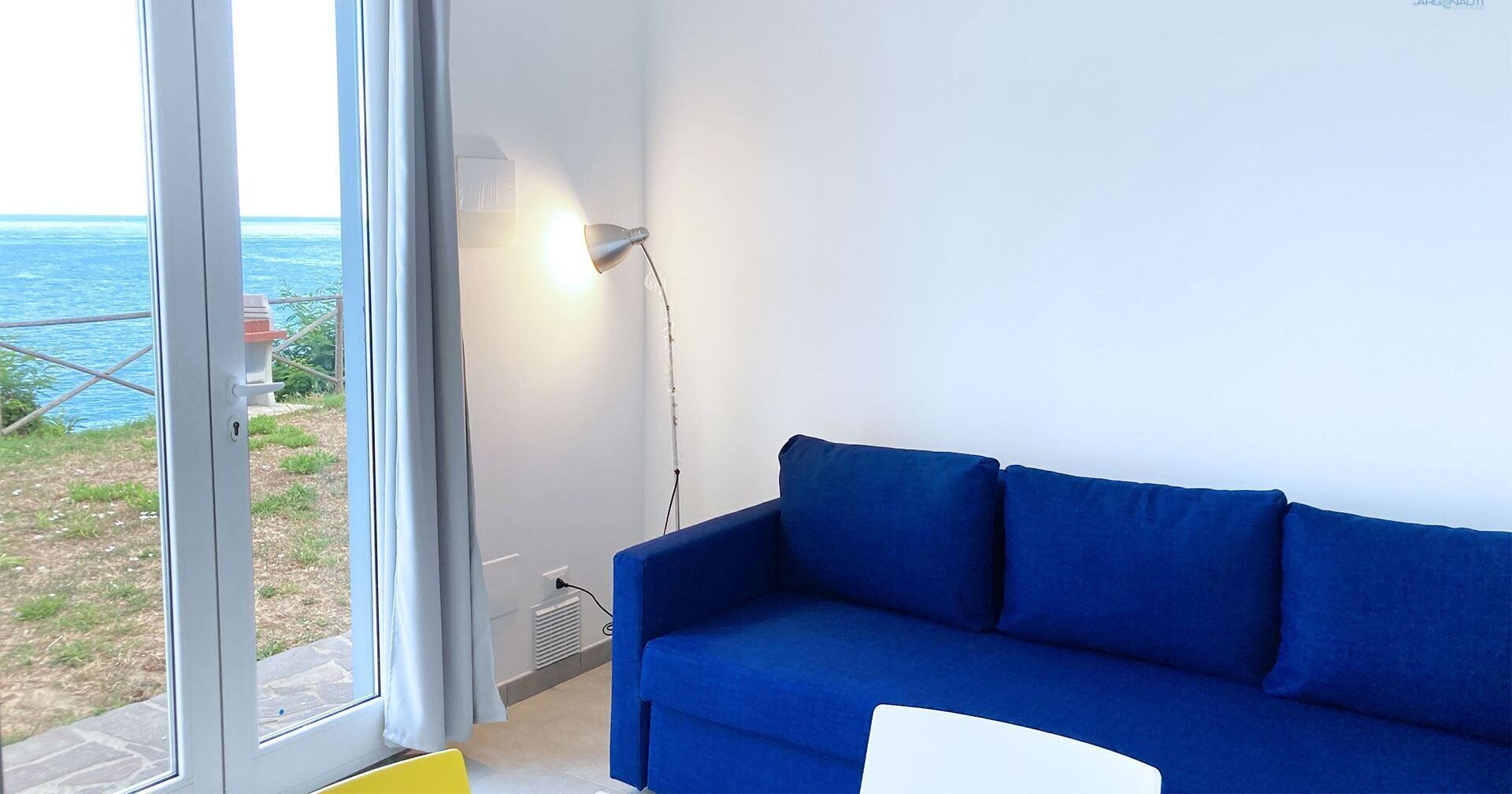 A room with a blue sofa, a lamp, and a view of the sea through a window.