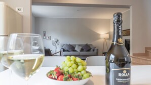 Ein Wohnzimmer mit grauem Sofa, einem Ventilator und einem Tisch mit einer Flasche Bolla-Wein, einer Obstschale und Gläsern.