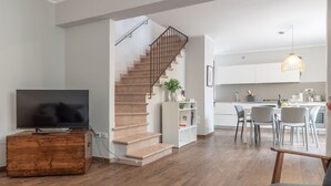 Ein modernes Wohnzimmer mit einem Holz-TV-Schrank, einer Treppe und einer Küchenzeile mit weißen Schränken und einem Esstisch.