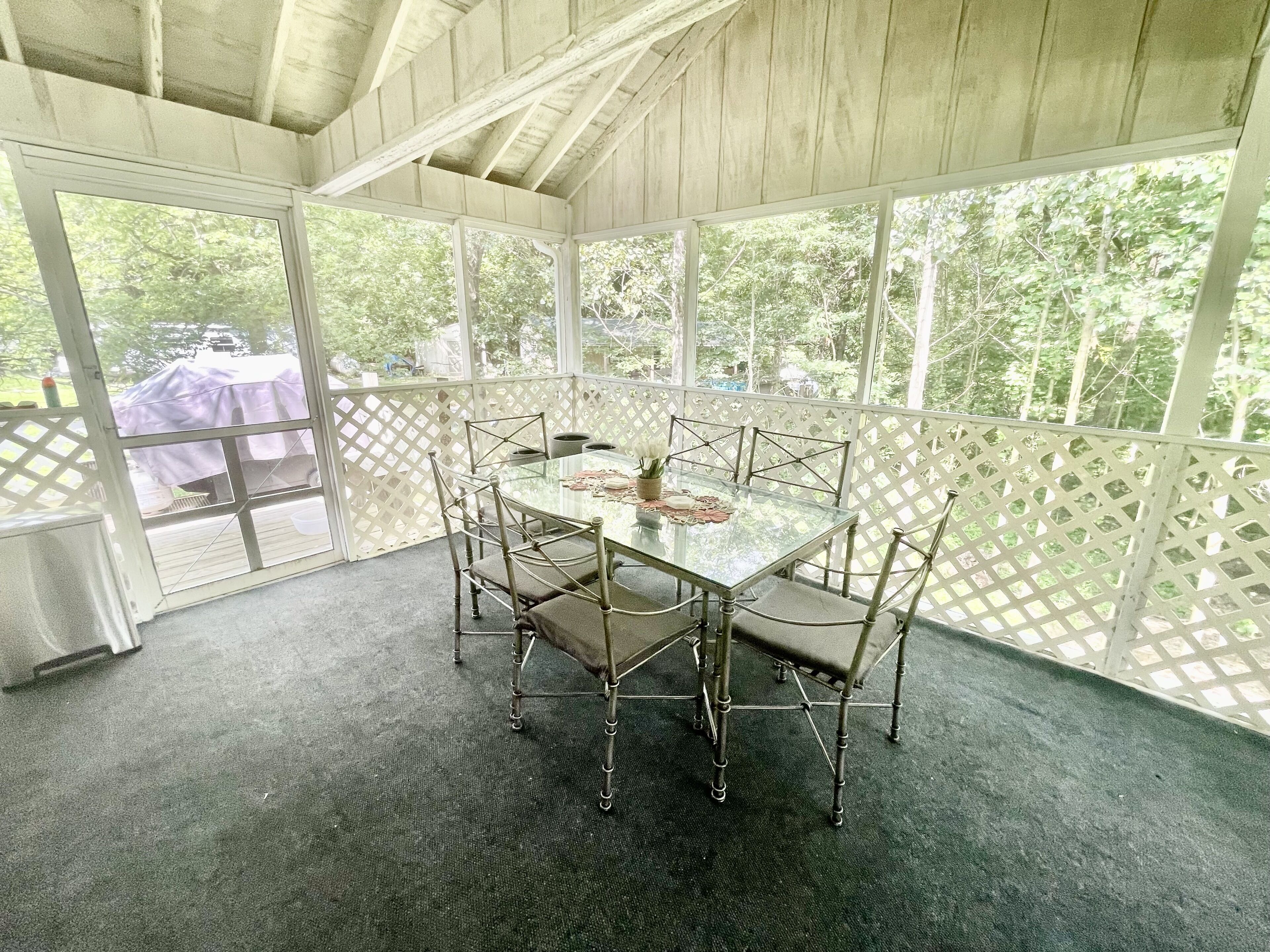 A covered outdoor dining area with a glass table and metal chairs, surrounded by a white lattice fence, and a view of greenery outside.