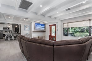 A modern living room with a large brown sectional sofa, a kitchen area with bar stools, and a television mounted on the wall.