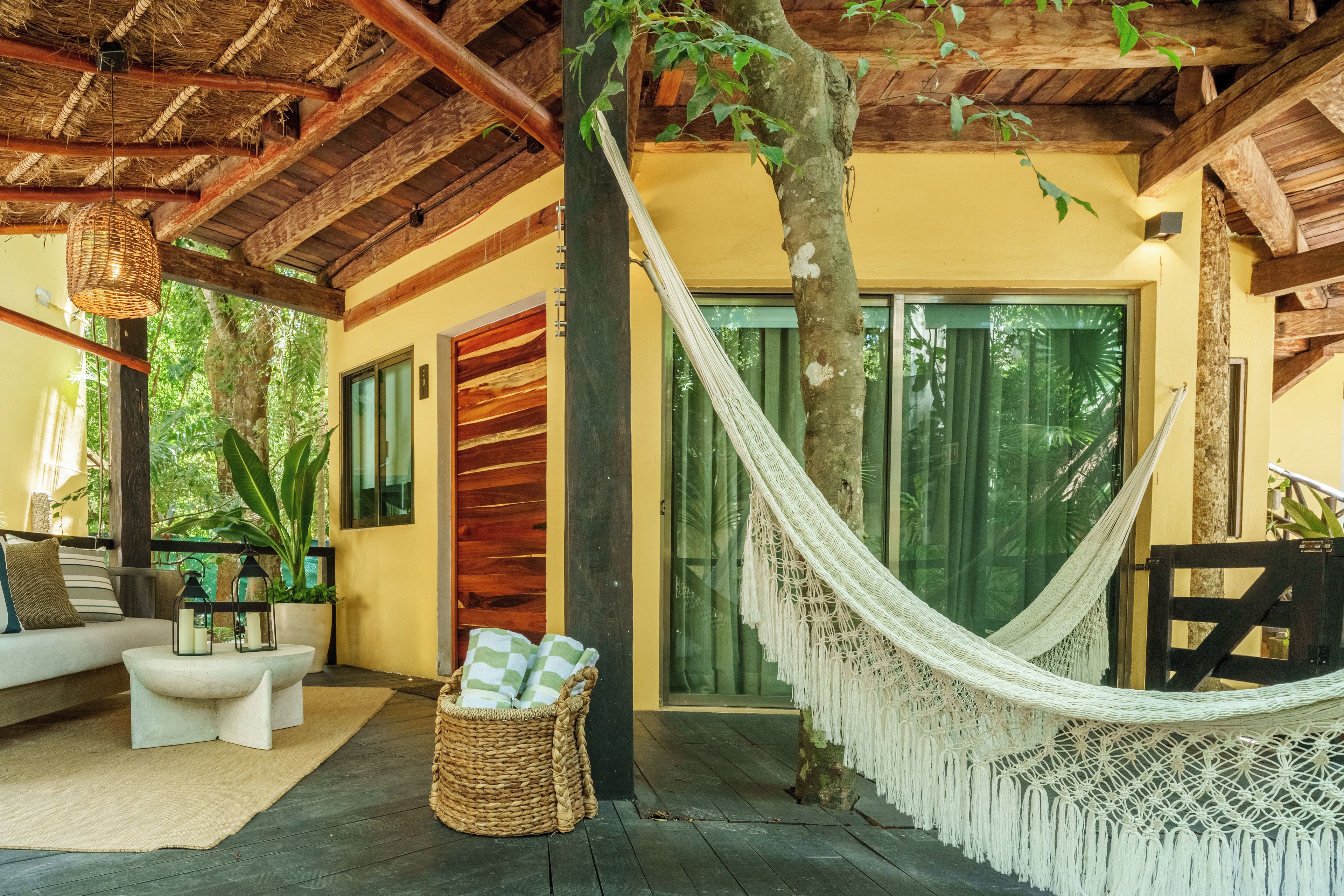 A hammock, a sofa, and a wicker basket in a covered outdoor area with a wooden ceiling and a yellow wall.