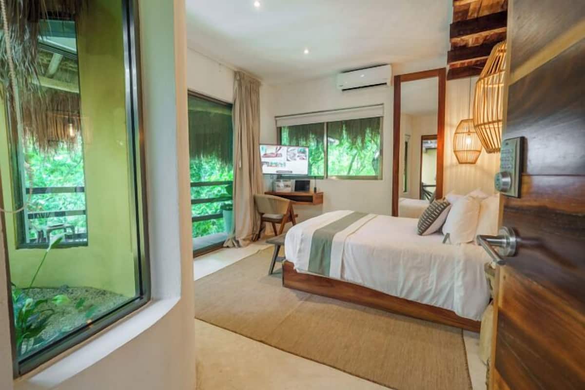 A bedroom with a bed, a desk, a TV, and a large window with a view of greenery.