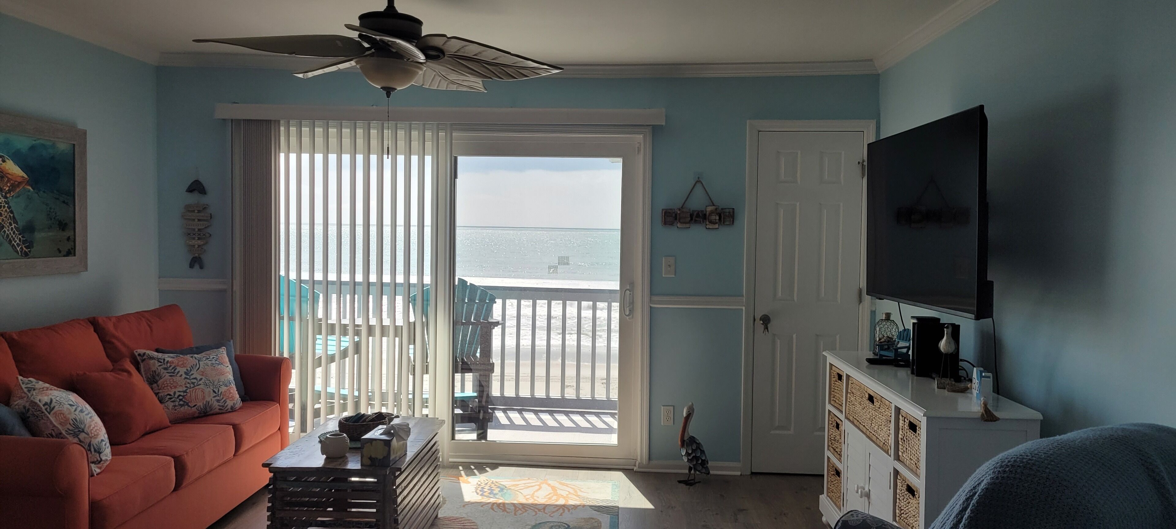 A living room with a sofa, a television, a ceiling fan, and a view of the beach through a sliding door.