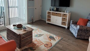 Una sala de estar con un ventilador de techo, un televisor, un armario blanco con cestas, un tapete estampado y un sofá azul con estampado.