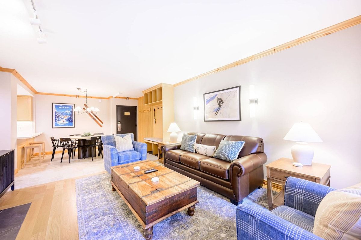 A living room with a brown leather sofa, a blue plaid sofa, a wooden coffee table, and a dining area with a table and chairs.