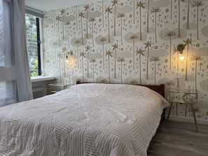 Une chambre avec un grand lit, une fenêtre avec des rideaux, un mur recouvert d’un papier peint à motifs naturels et deux lampes fixées au mur.