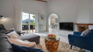 Un salon moderne avec un canapé, un fauteuil bleu et une table basse en verre sur laquelle sont disposés des fruits. Un téléviseur est fixé au mur, et il y a une cheminée.