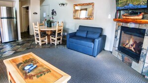 Ein Wohnzimmer mit Kamin, einer Couch, einem Esstisch mit Stühlen, einem Couchtisch und einer Küchenzeile inklusive Kühlschrank.