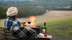 Eine Person sitzt auf einer Bank, im Hintergrund brennt ein Feuer, dazu ein Glas Wein und ein Teller mit Käse und Trauben.