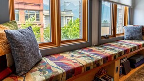 Asiento junto a la ventana con un cojín de colores, vista de un edificio y una estantería con libros.