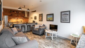 Una sala de estar moderna con un sofá gris, una mesita y una cocina con pared de ladrillo y taburetes de bar.