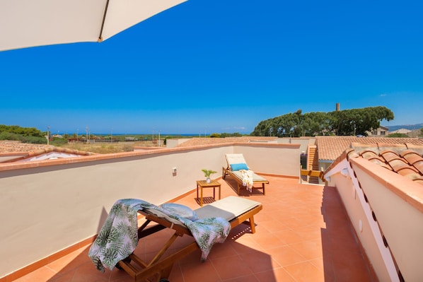 A rooftop terrace with a clear view of the sea, a lounge chair, a small table with a plant, and a tiled floor.