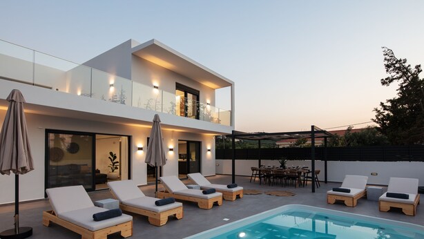 A modern outdoor pool area with lounge chairs and umbrellas, a dining set, and a two-story building with glass railings and lighting. - Horizon Villa Oasis, Walk to Platanias Beach! (Chania)