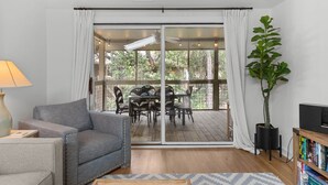 Una sala de estar con sofá, una mesa de centro, un mueble de televisión con libros, una planta en maceta y un ventanal corredero que da a un patio exterior con una mesa y sillas.