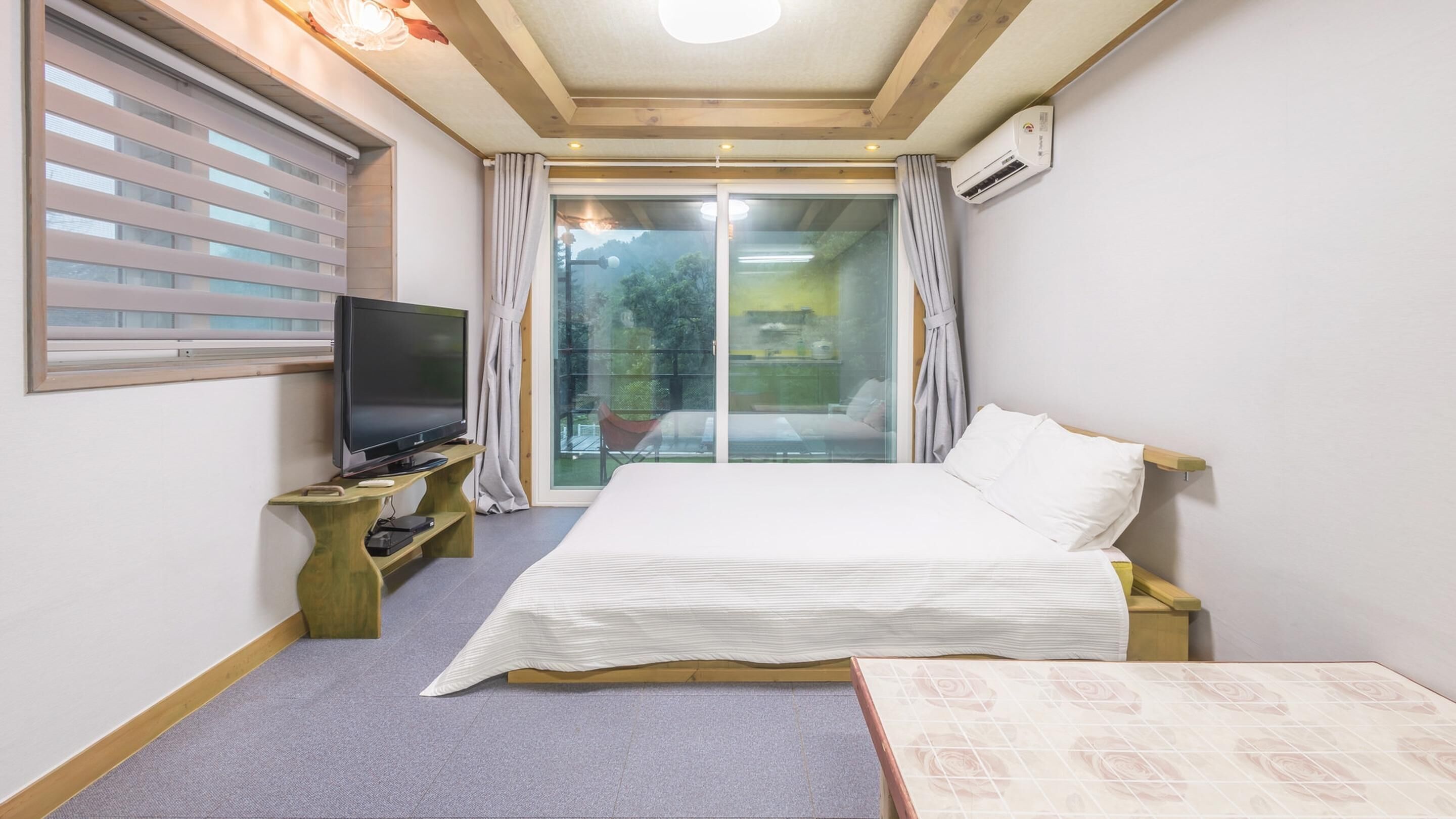 Een hotelkamer met een bed, een televisie op een houten standaard, een raam met zonwering en een airconditioner.