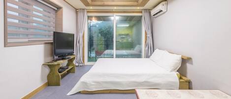 Een hotelkamer met een bed, een televisie op een houten standaard, een raam met zonwering en een airconditioner.
