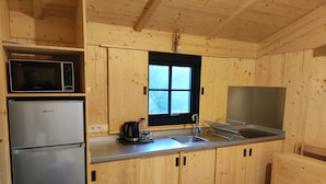 A compact kitchen with wooden cabinets, a stainless steel refrigerator, a microwave, a sink with a drying rack, and a window above the sink. - LE JARDIN SAINT-GERMAIN Lodges & Events (Plogastel-Saint-Germain)