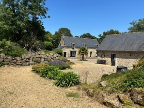 Un bâtiment en pierre doté d’un toit en pente, entouré d’une cour recouverte de gravier et agrémentée de diverses plantes.
