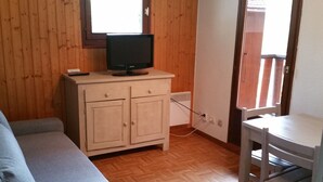Ein Zimmer mit Holzboden, einem grauen Sofa, einem kleinen weißen Tisch und einem Fernseher auf einer Holzkommode.