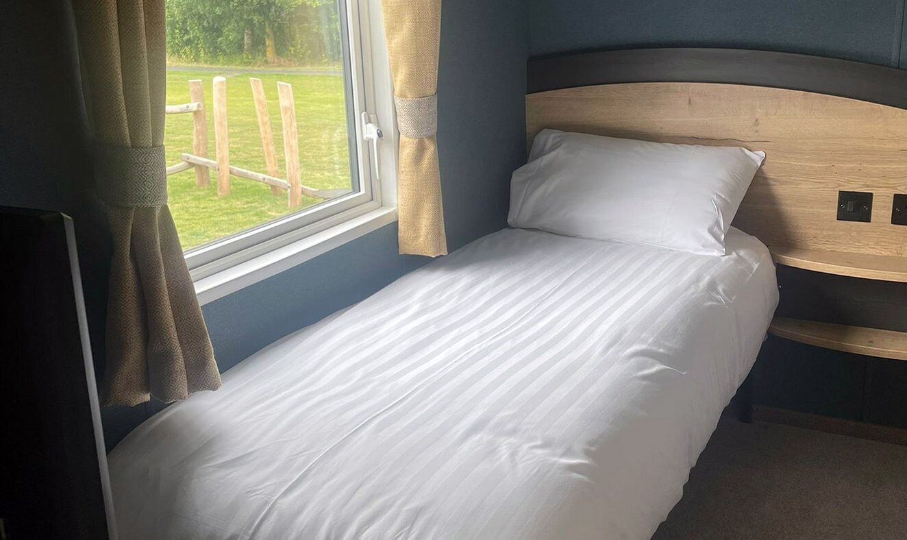 A single bed with a wooden headboard, a window with curtains, and a view of a grassy area outside.