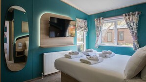 Ein Schlafzimmer mit türkisfarbenen Wänden, einem großen Bett mit weißer Bettwäsche, einem flachbildfernseher, der über dem Bett montiert ist, einem Spiegel und einem Fenster mit gemusterten Vorhängen.