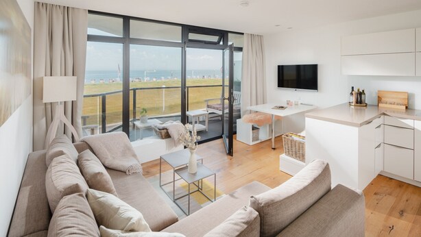 Un salon moderne avec un grand canapé, une table à manger et un téléviseur. De grandes fenêtres offrent une vue sur la mer.