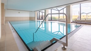 Une piscine intérieure à l’eau d’un bleu limpide, avec un escalier en métal et une vue sur la plage grâce à de grandes fenêtres.