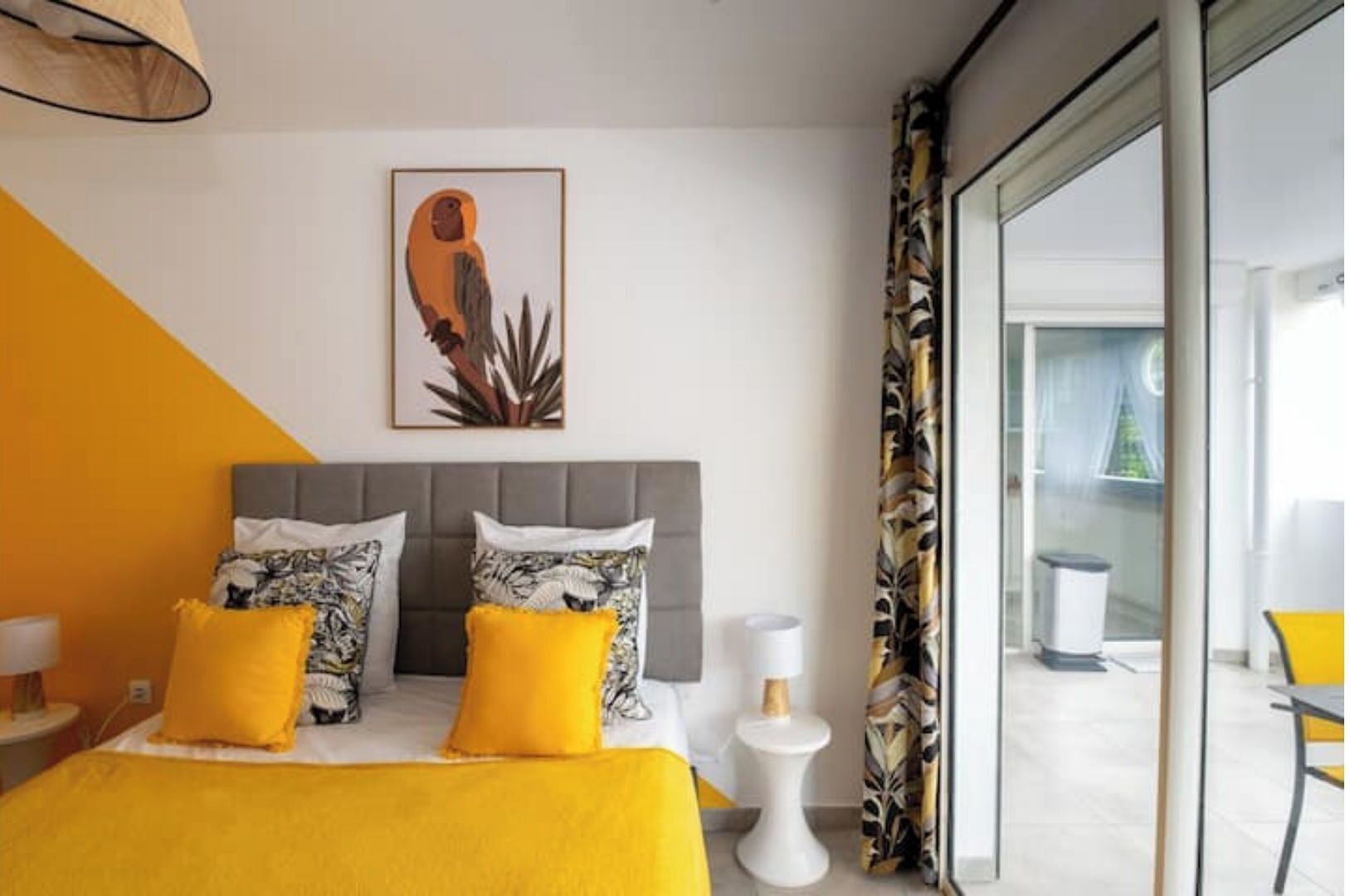 A modern hotel room with a bed, a bedside table, a lamp, a framed artwork, and a view of the outside.