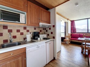 Kompakti keittiö, jossa on puukalusteet, Bosch-astianpesukone, mikroaaltouuni ja kahvinkeitin. Keittiössä on myös ruokailutila, jossa on punainen sohva ja pöytä.