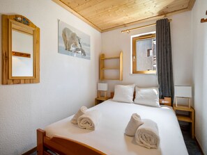 A hotel room with a wooden ceiling, a bed with white linens, two rolled towels, a painting of a polar bear, a window with a curtain, and a wooden nightstand.