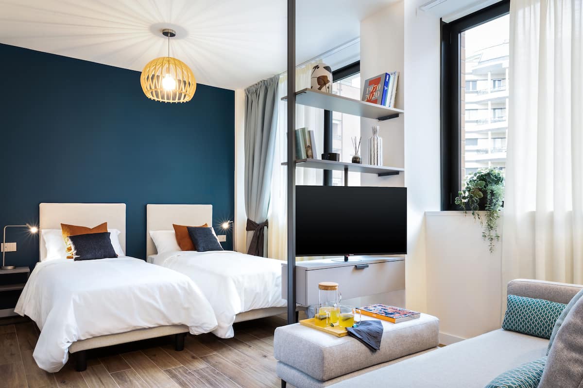 A modern hotel room with two beds, a flat-screen TV, a sofa, and a dining area with a tray of breakfast items.