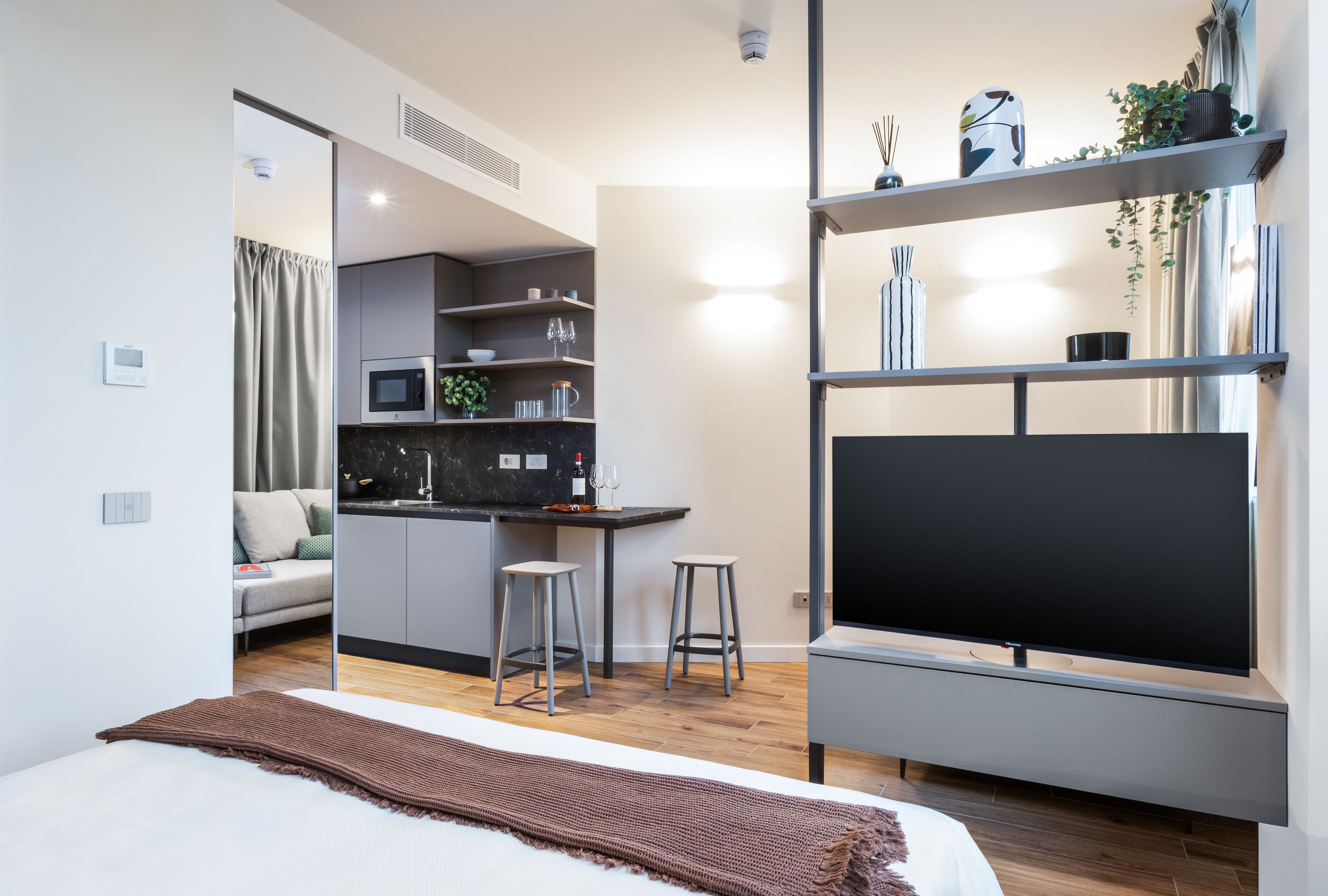 Quarto de hotel moderno com cama, televisão, cozinha compacta e área de estar.