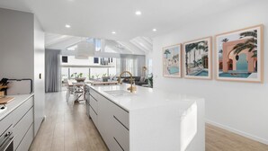 A modern kitchen with a central island, white cabinetry, and a dining area with a grey sofa. - Gone Coastal - Kingscliff (Kingscliff)