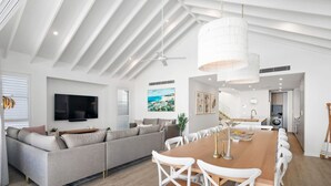 A modern living room with a large sofa, a dining table with white chairs, a TV mounted on the wall, and a ceiling fan. - Gone Coastal - Kingscliff (Kingscliff)
