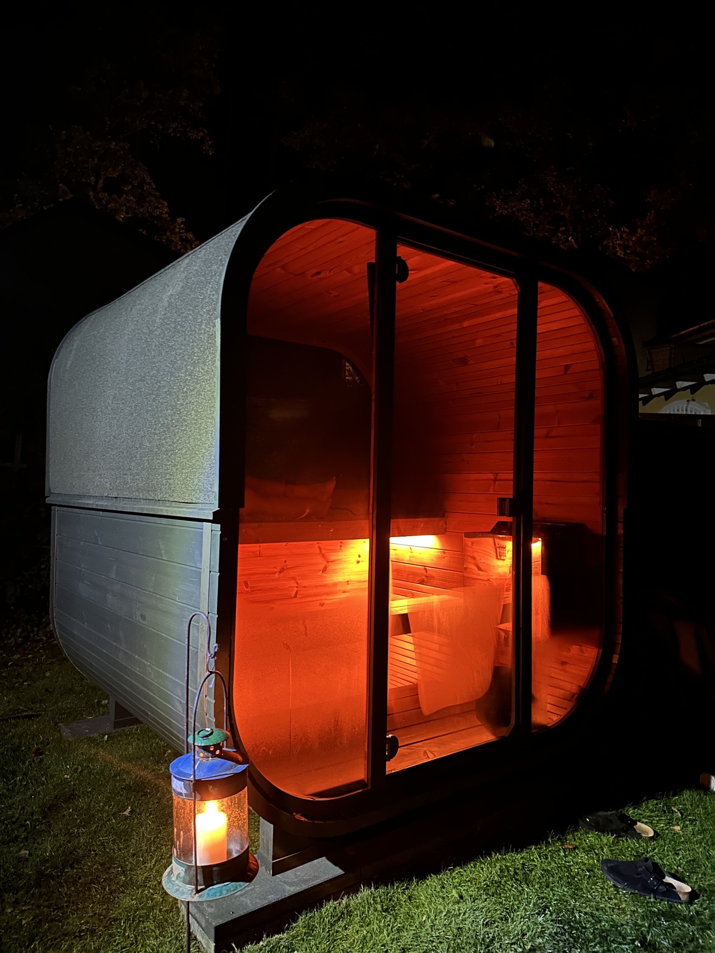A small, enclosed wooden structure with a glass door, a lit lantern outside, and a pair of shoes on the grass.