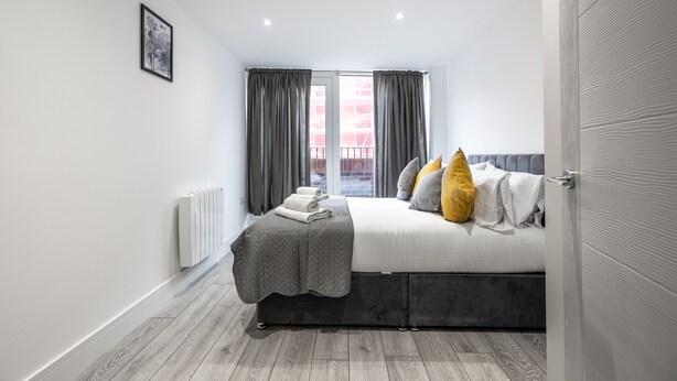 A modern bedroom with a bed, grey curtains, a radiator, and a window with a view. - Stunning 2-bed Apartment in Camberley (Camberley)