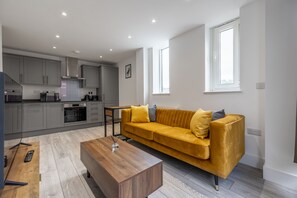 A modern apartment with a kitchen, a yellow sofa, a wooden coffee table, and a large window. - Stunning 2-bed Apartment in Camberley (Camberley)