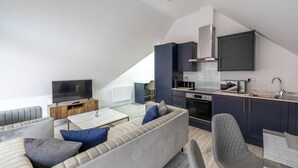 A modern loft apartment with a living area featuring a sofa and a flat-screen TV, a kitchen with dark cabinetry and a countertop, and a dining area with a table and chairs. - Impeccable 2-bed Apartment in Camberley (Camberley)