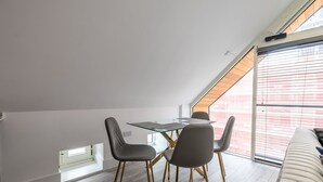 A modern dining area with a glass table and chairs, a sloped ceiling, and a view of a red building through large windows. - Impeccable 2-bed Apartment in Camberley (Camberley)