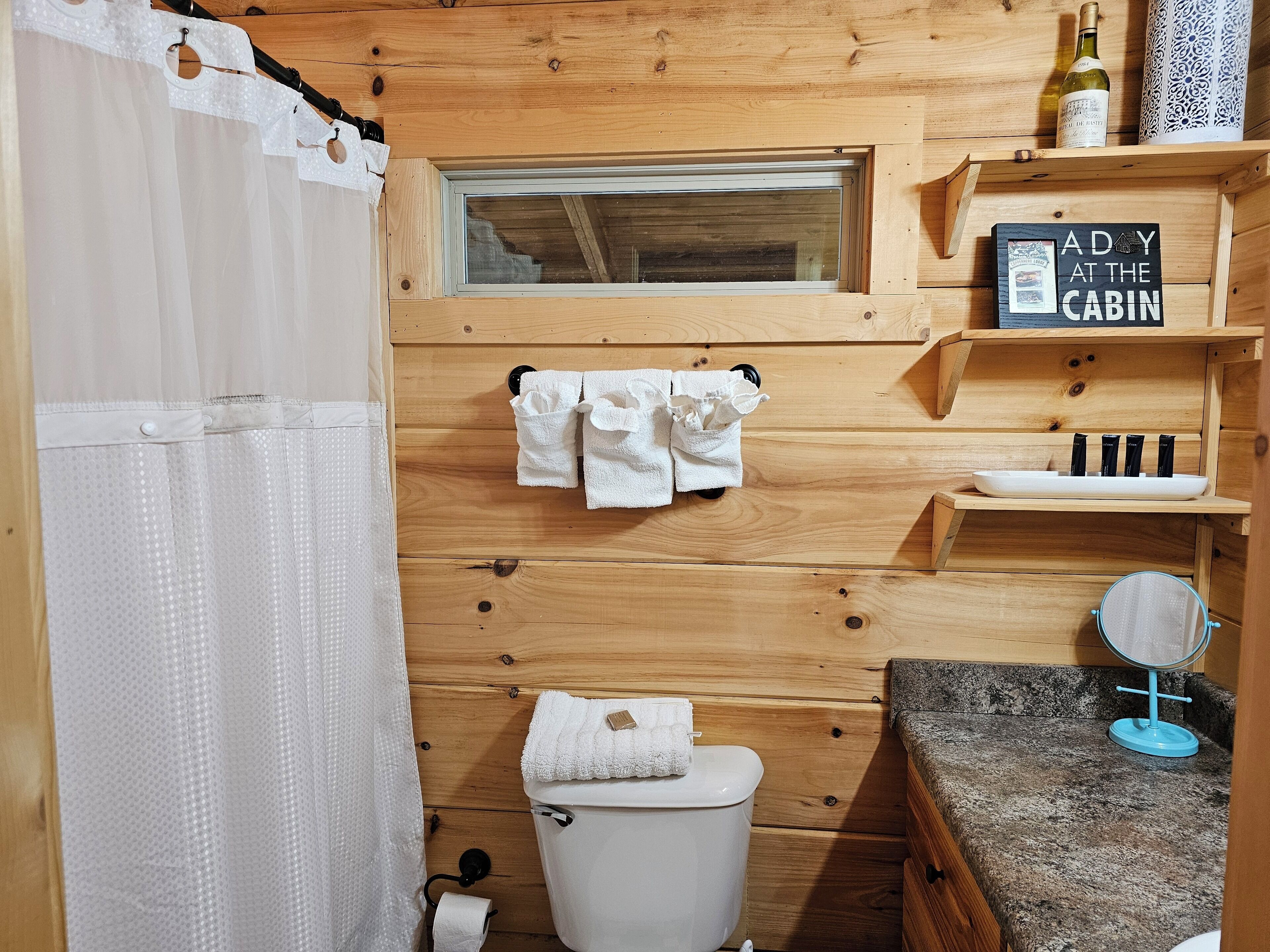 A wooden cabin bathroom with a toilet, a window, a shelf with a sign, and a towel rack.