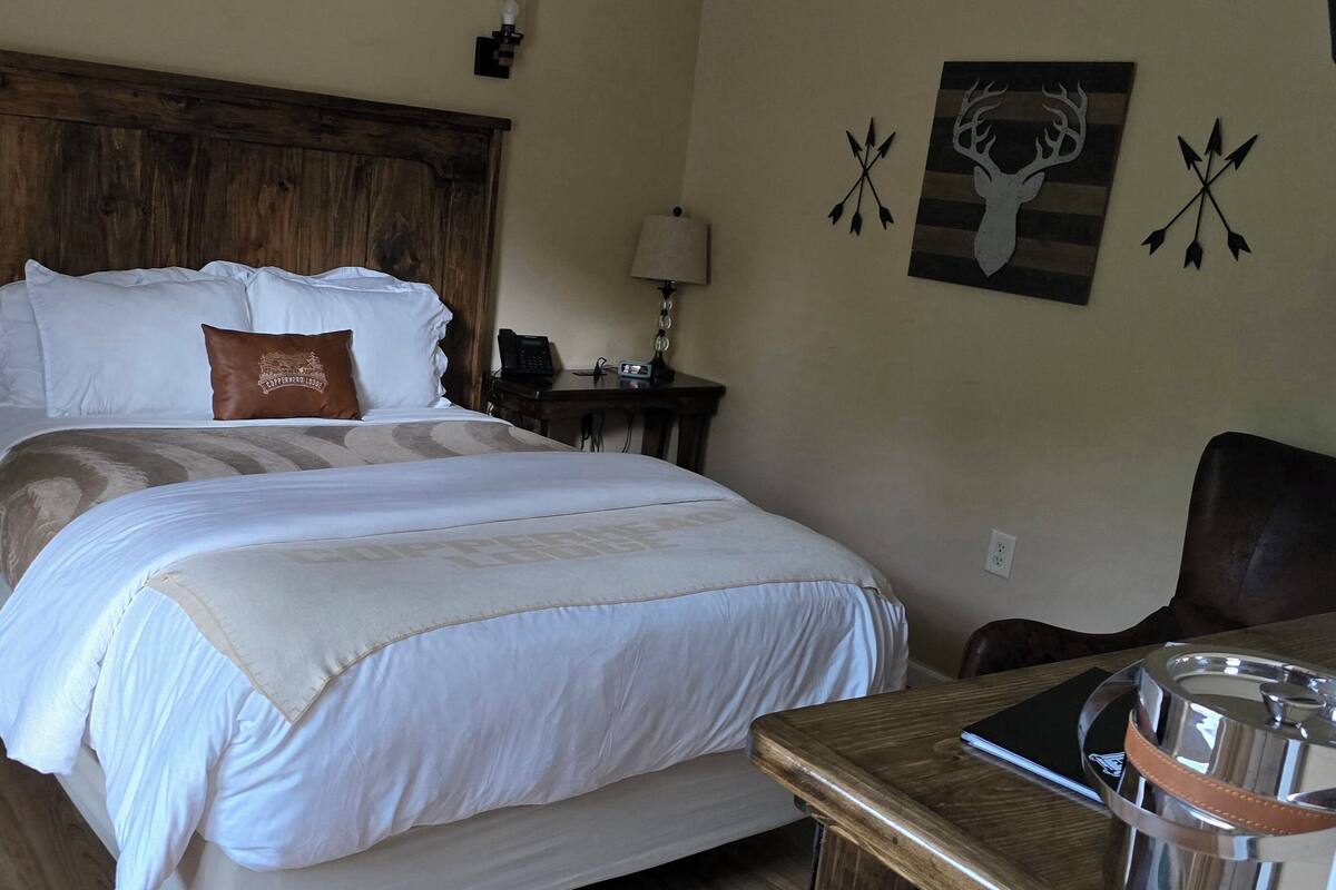 A neatly made bed with a white and beige bedspread, a brown pillow, a wooden headboard, a bedside table with a lamp, a phone, and a wall with a deer head and arrow decorations.
