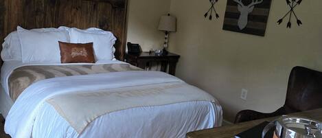 Una cama bien hecha con colcha blanca y color crema, almohada marrón, cabecero de madera, mesita de noche con lámpara, teléfono y pared decorada con motivos de cabeza de ciervo y flechas.