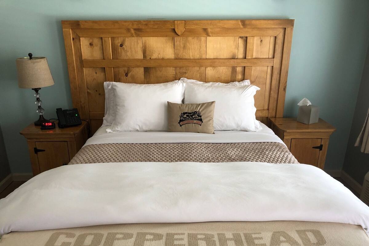 A neatly made bed with a wooden headboard, two white pillows, a beige throw blanket, and a pillow with a logo. There is a bedside table with a lamp, a phone, and a tissue box.