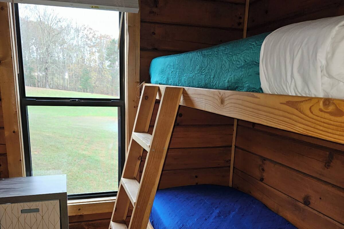 A bunk bed room with a wooden ladder, a small desk, and a window with a view of a grassy area.